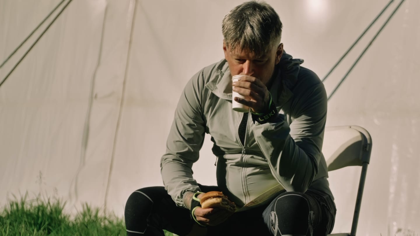 A walker at a night checkpoint on the Cateran Yomp, sitting in a lit tent with a paper cup in one hand and a burger in the other, eyes down.