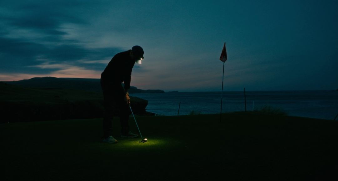 A golfer stands over a putt by head torch at dusk on the ninth green at Durness, the sea and the last light behind him.