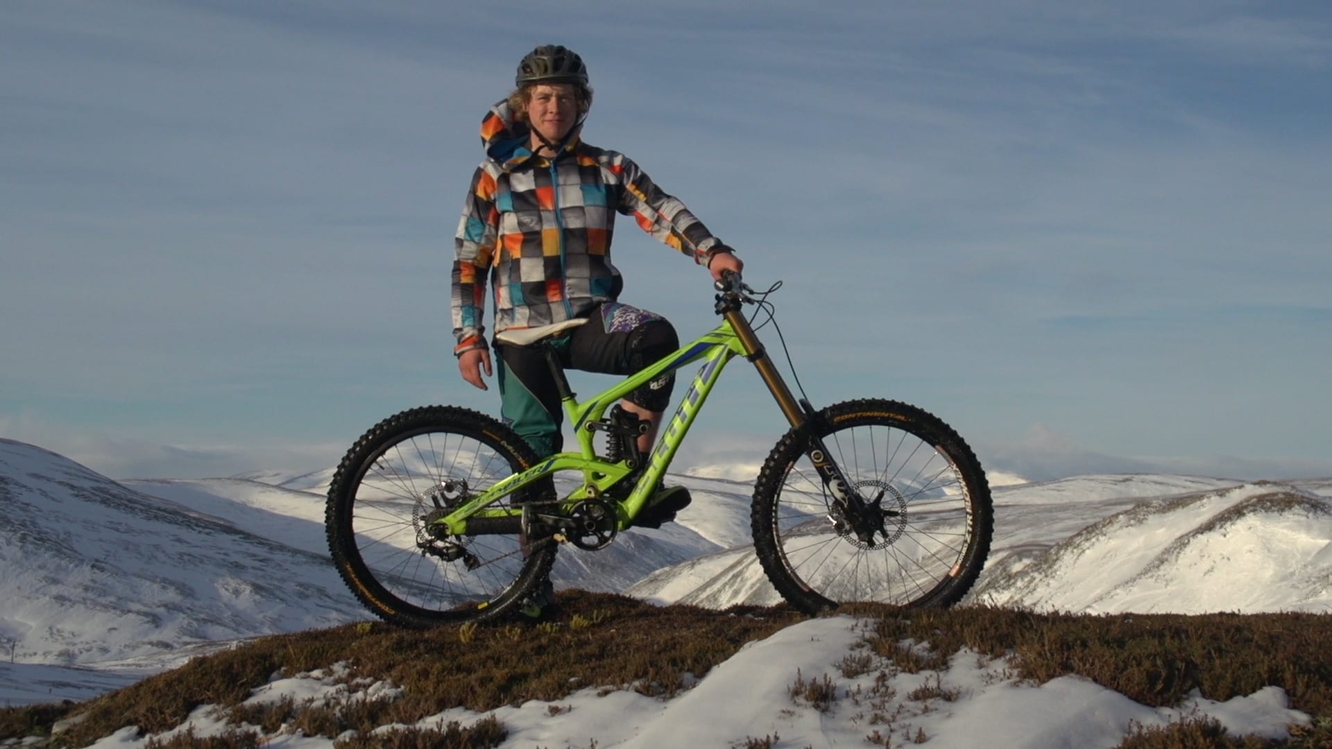 A young mountain biker stands with his bike on a snowy Scottish hilltop under a clear sky.
