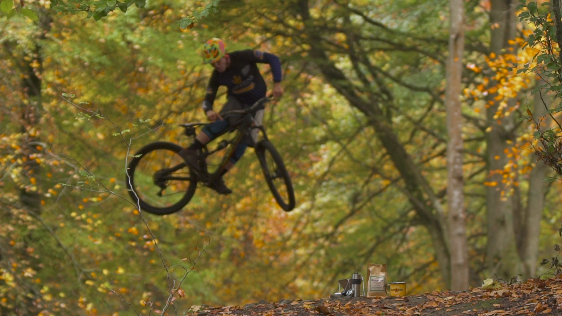 A mountain biker catches air in an autumn wood, with a coffee brand cafetiere, bag and mug staged in the foreground.
