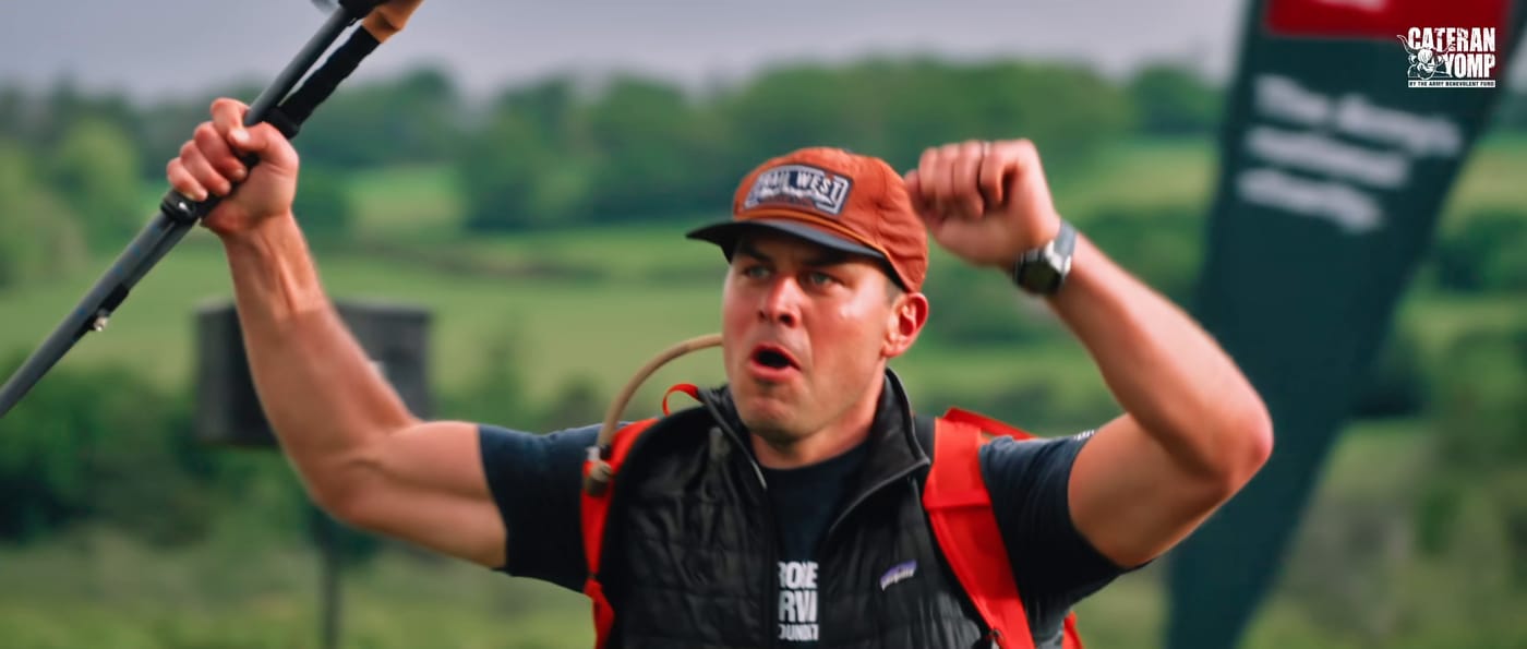 A walker at the finish with both arms raised and a pole in one hand, mouth open, shouting.