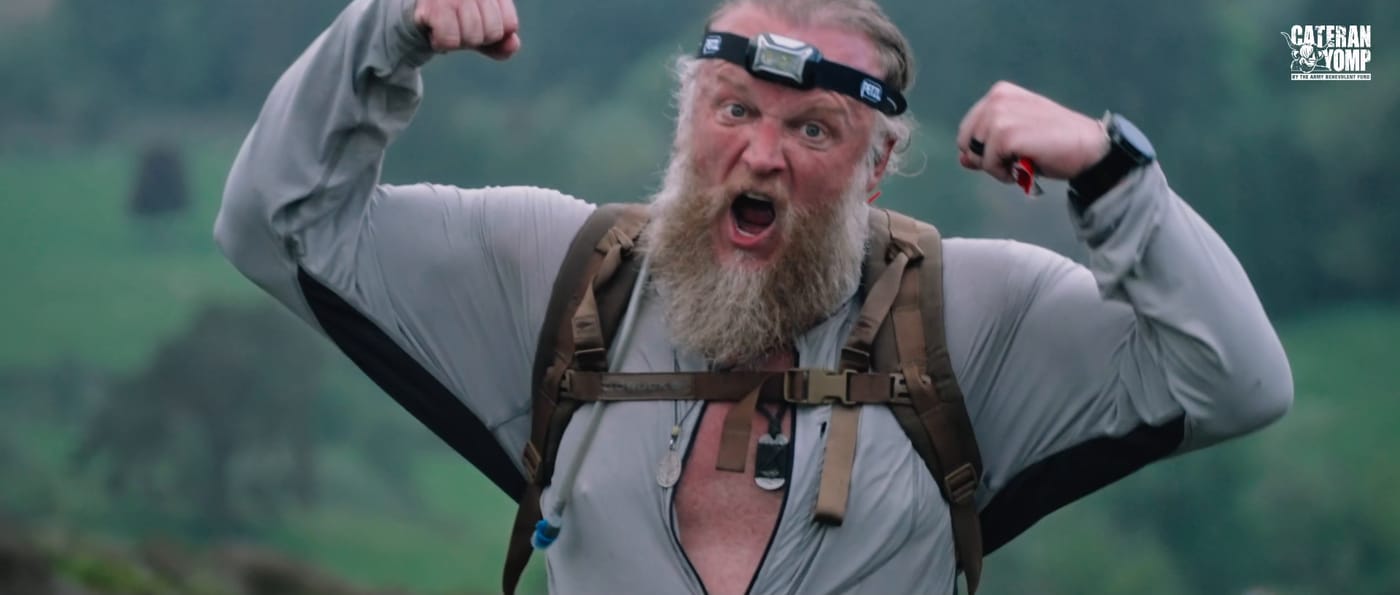 A bearded walker with a head torch on his forehead roaring and flexing both arms at the top of a climb.