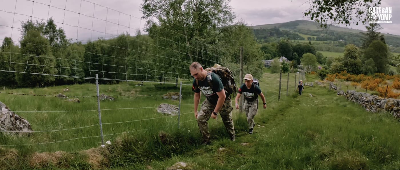 Two walkers climbing a grass track beside a wire fence, a third further up the hill ahead of them, green farmland beyond.
