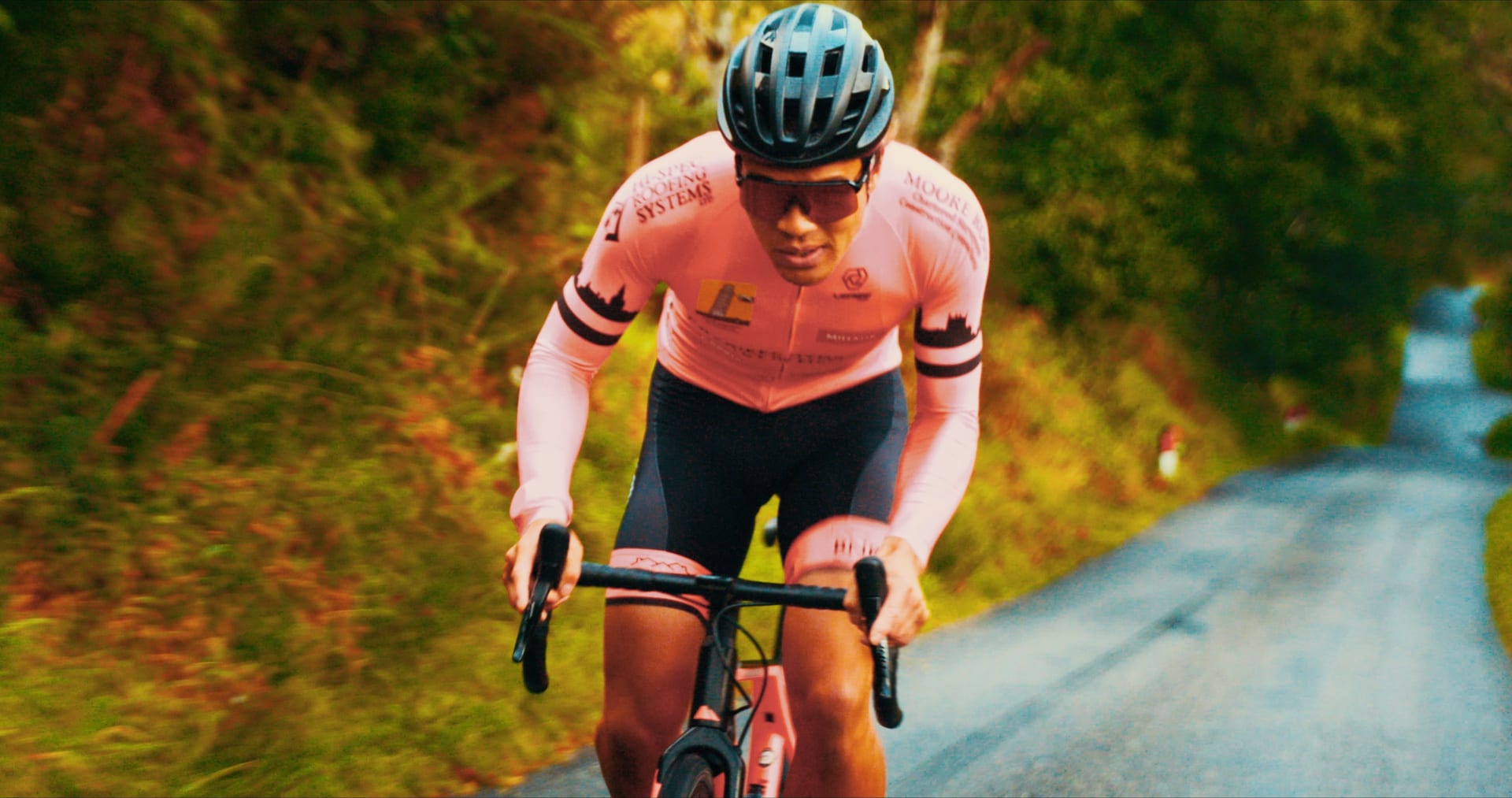 A road cyclist in race kit pushes hard along a country lane in low autumn light.