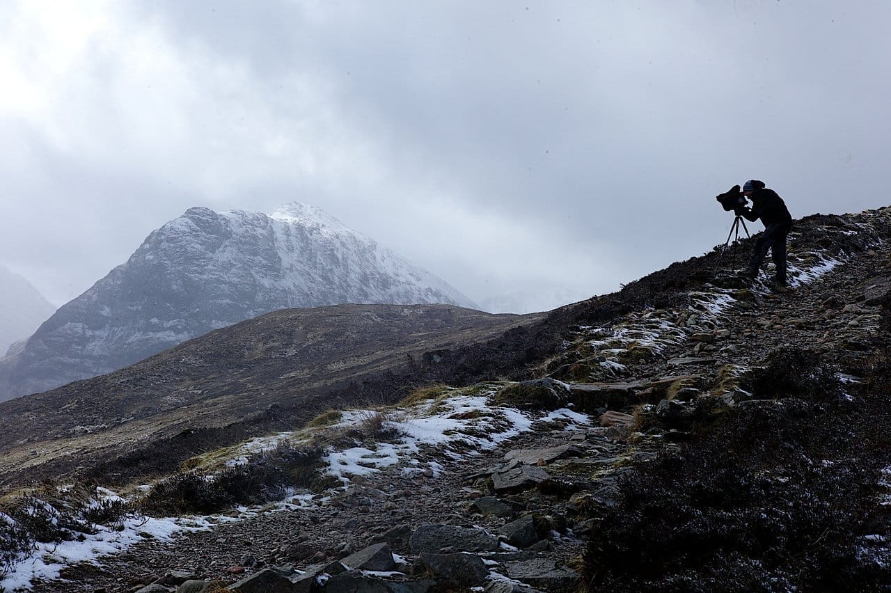 A lone filmmaker on a snowy Highland ridge, camera trained on a cloud covered peak.