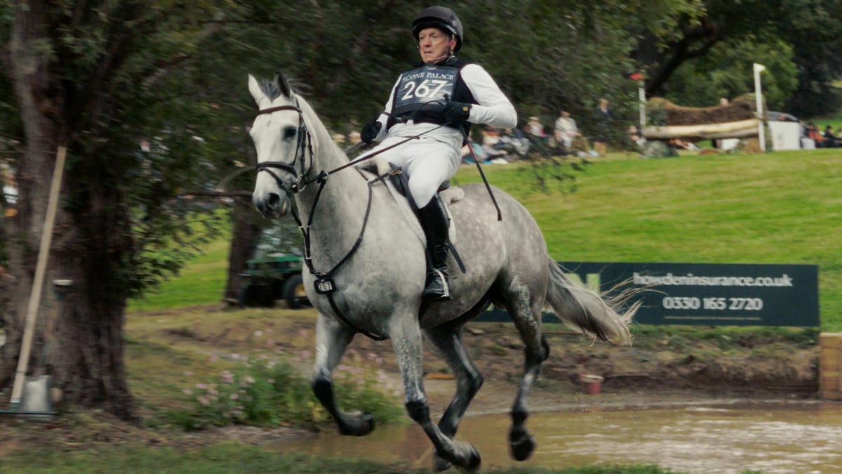 A rider gallops a grey horse through water at a cross country event, eyes fixed on the line ahead.