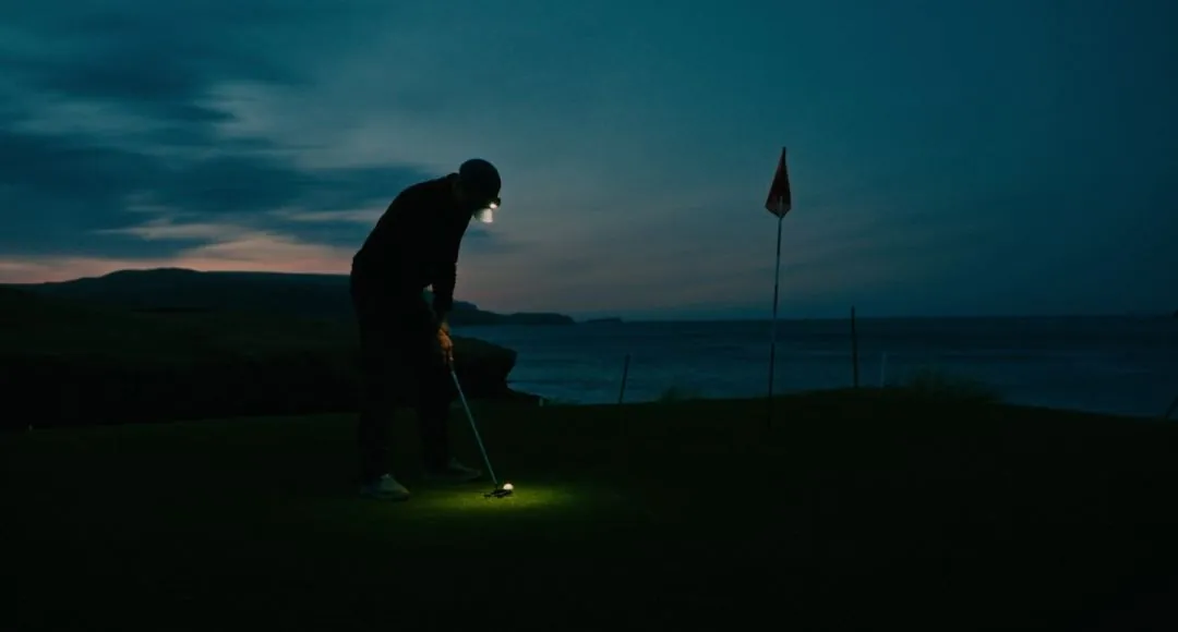 A golfer stands over a putt by head torch at dusk on the ninth green at Durness, the sea and the last light behind him.