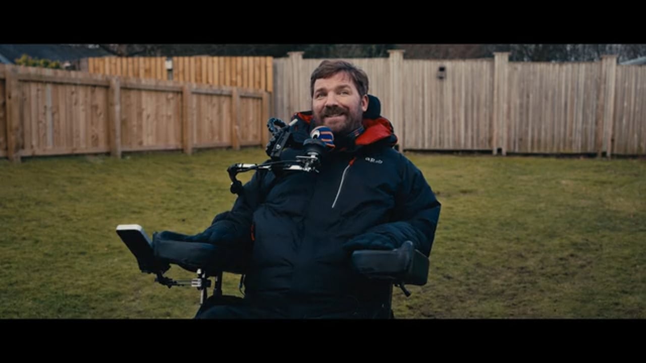 Danny Mavor smiles in his powered wheelchair in a garden, mid interview.